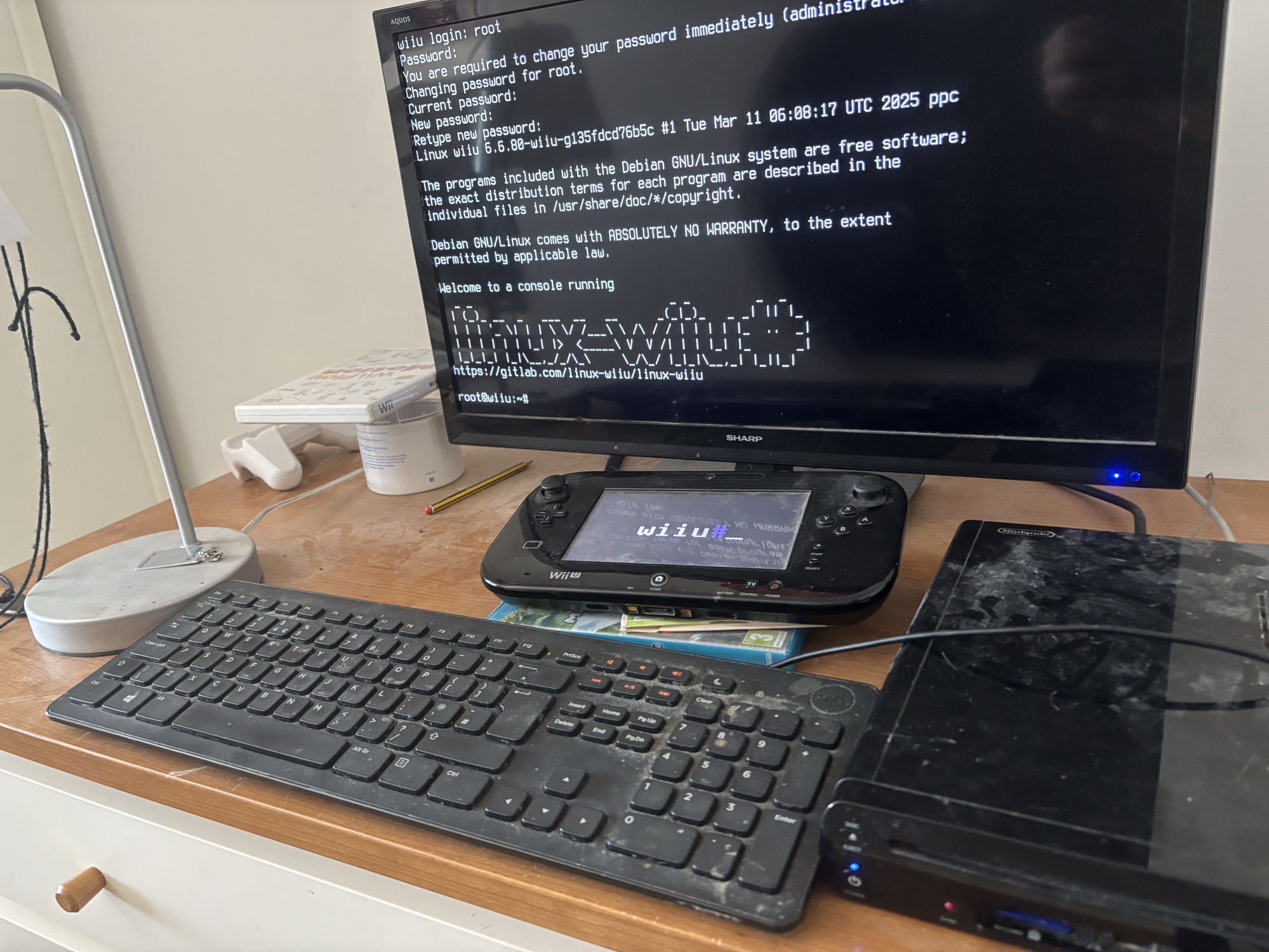 The setup showing a Sharp TV displaying a successful Debian Linux boot screen on a Wii U console. The terminal shows login prompts and system information including ‘Linux wiiu 5.8.0-wiiu-gba50f3cd76b5c’ and welcome messages. A black Wii U GamePad is visible on the wooden desk alongside a black wireless keyboard, with the Wii U console positioned to the right. The setup demonstrates homebrew Linux installation on Nintendo’s gaming hardware.